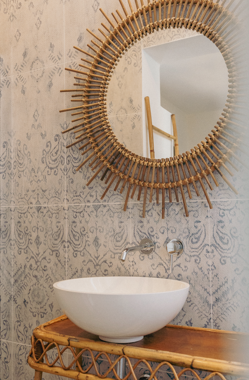 Round rattan mirror above a white sink on a wooden vanity with patterned wallpaper.