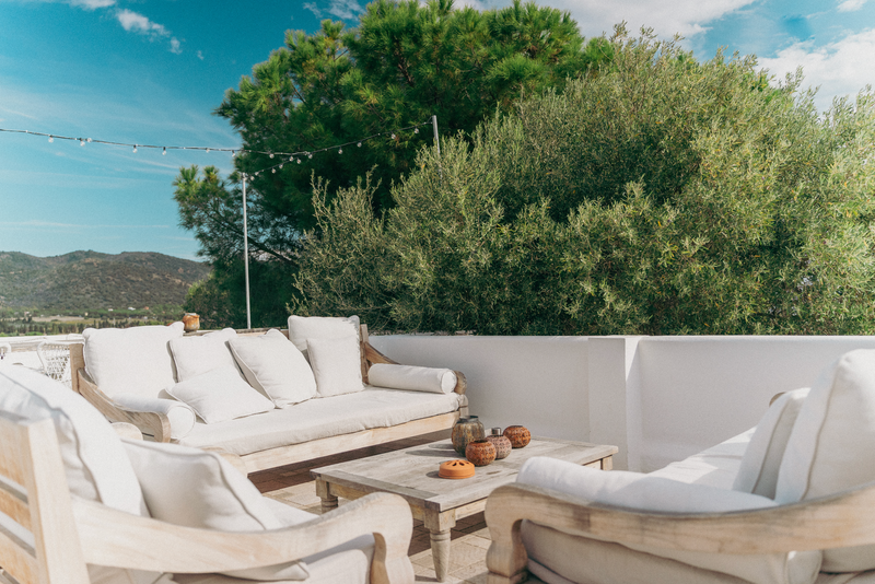 Outdoor seating area with white cushions and wooden furniture against a backdrop of green trees and mountains.