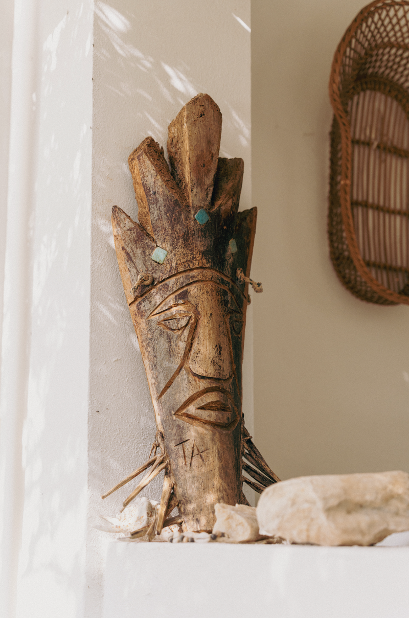 Wooden totem-like sculpture with face and feathers on a white surface